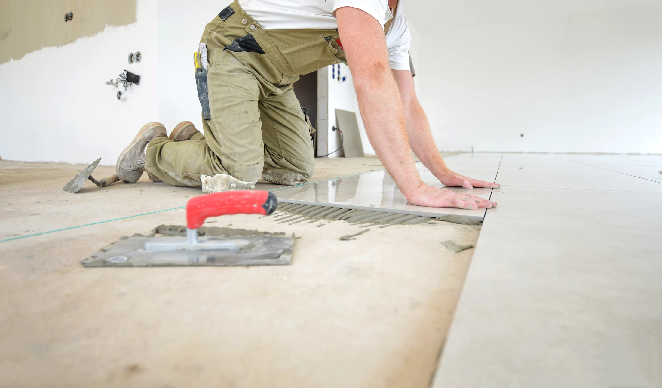 Artisan carreleur à genoux posant du carrelage dans une pièce en rénovation, murs blancs partiellement peints
