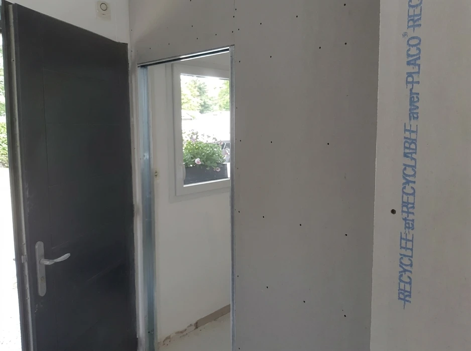 Chantier intérieur en cours, pièce vide avec porte d'entrée métallique anthracite et fenêtre avec jardinière