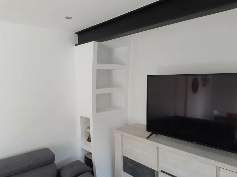 Salon moderne avec meuble TV en bois clair, téléviseur LG grand écran sur pied, bibliothèque encastrée en placo peint blanc avec niches et étagères